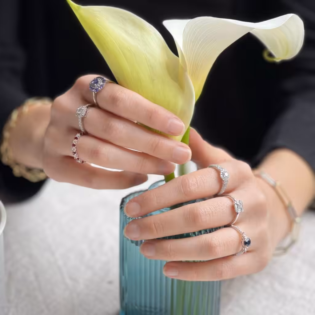 Dos manos con distintos anillos sosteniendo una cala blanca junto a un florero.