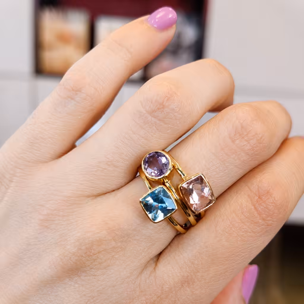 Mano con anillos contemporáneos de piedras celeste, violeta y rosada en oro.